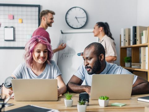Zwei Personen sitzen im Büro vor ihren Laptops, im Hintergrund stehen zwei Menschen vor einem Flipchart